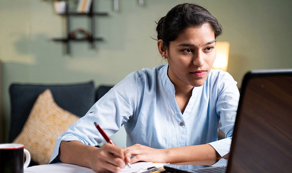 Young business woman writing down notes by looking laptop - concept of employee or student online training at home. Young business woman writing down notes by looking laptop - concept of employee or student online training at home
